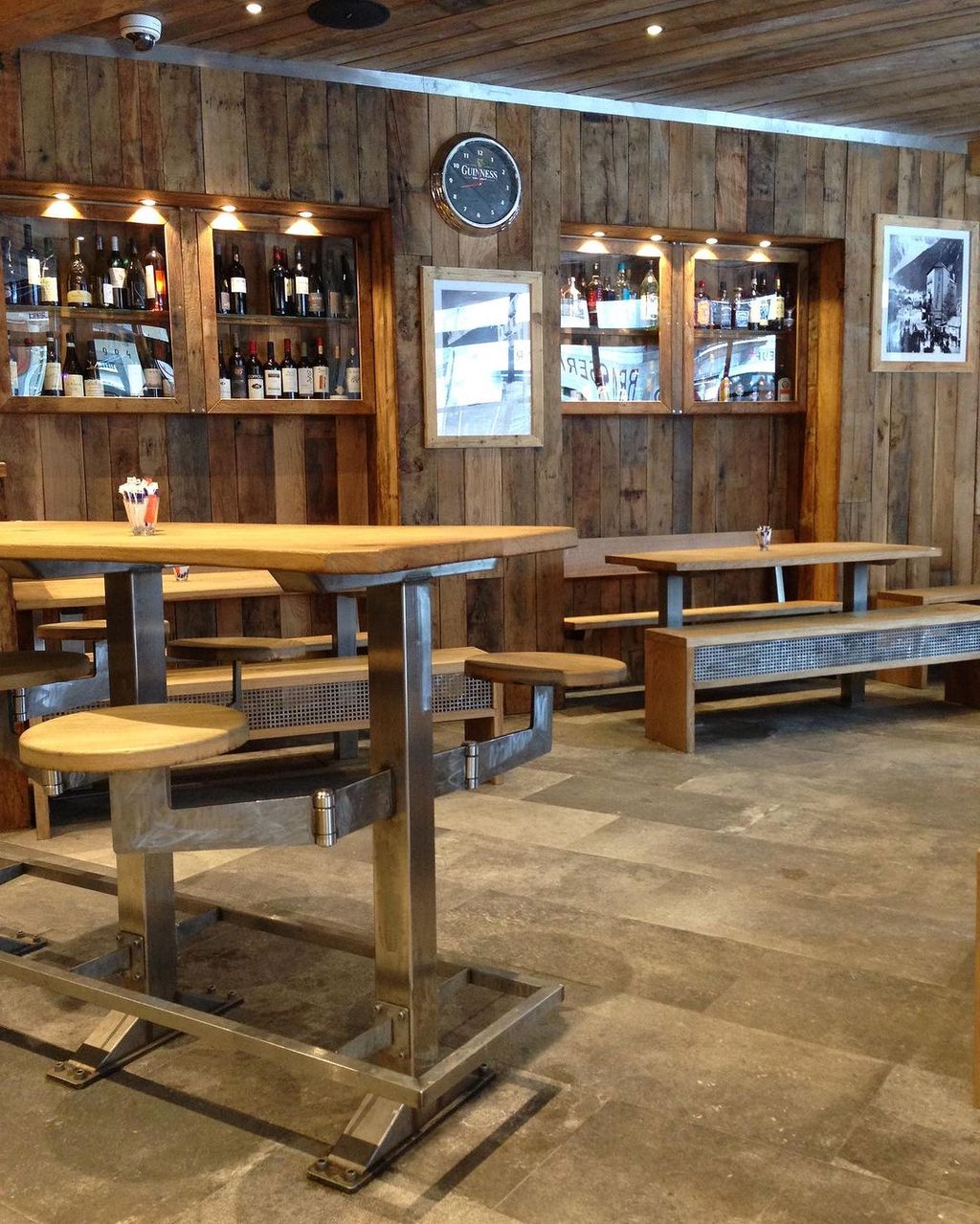 Cafe tables with wooden tops and metal legs, with shelves of wine in wooden cabinets embedded in the wall behind.