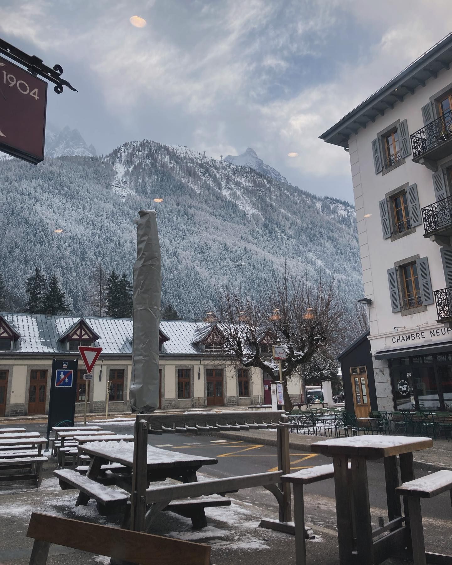 A sidewalk with 9 tables and benches that has snow on them, in front of a steep snow cowered forest with steep dark mountains peaks ricing into the clouds above them.