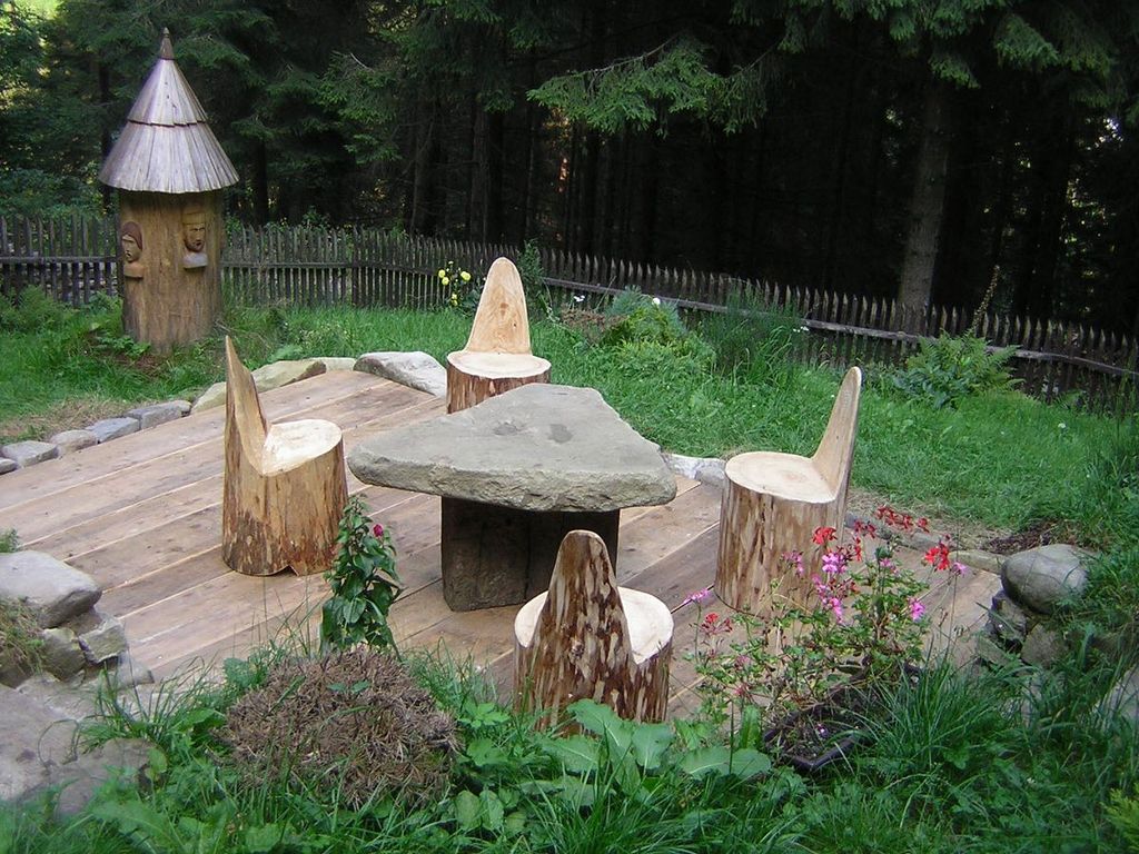 A lush green garden in a forest, a seating area with 4 chairs made out of big logs, around a table made out of two rocks balancing on top of each others.